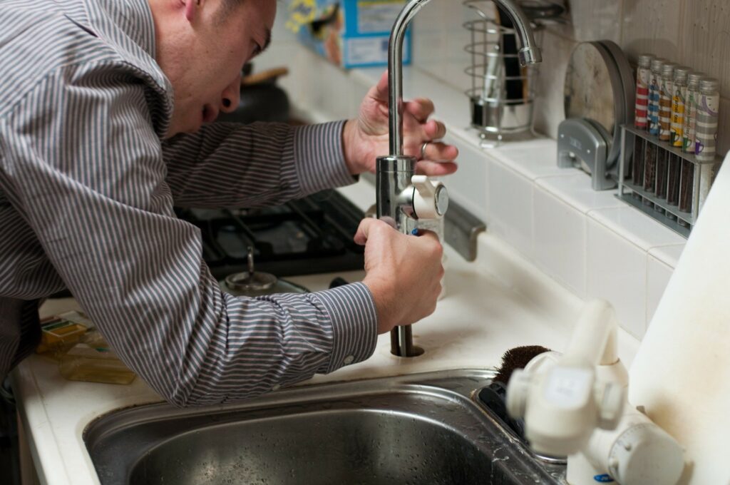 Man in a striped shirt repairing a kitchen faucet under the sink with his hands on the faucet valve.